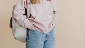 A teenager in a casual pink hoodie and jeans carrying a backpack, standing against a neutral background.