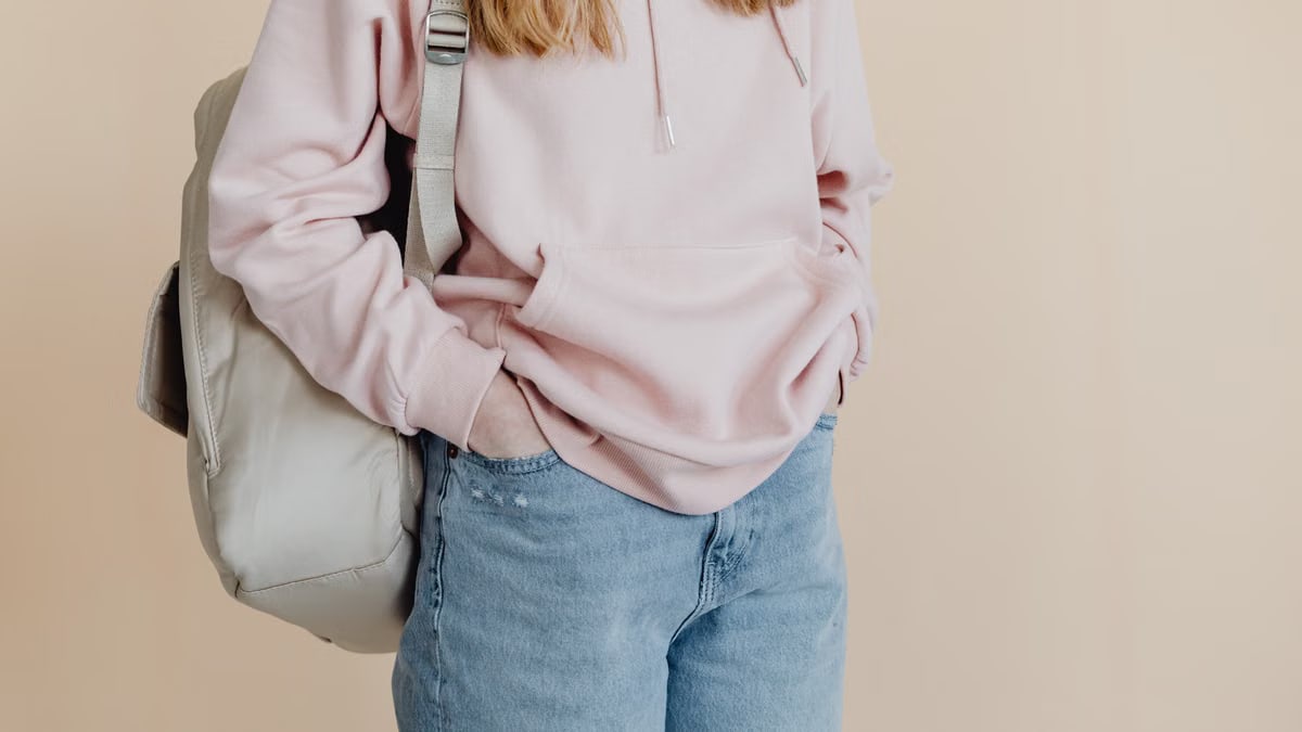 A teenager in a casual pink hoodie and jeans carrying a backpack, standing against a neutral background.